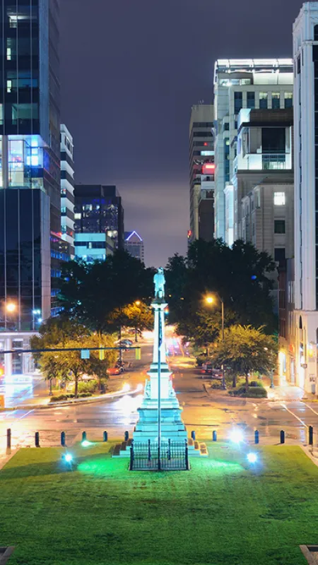 Nighttime cityscape with illuminated monument in green park surrounded by tall buildings and wet streets.