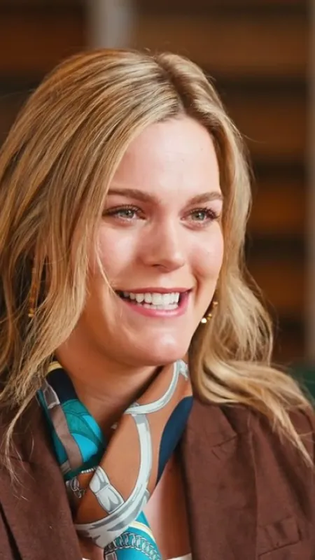 Smiling blonde woman wearing a brown blazer and patterned scarf in a warmly lit indoor setting