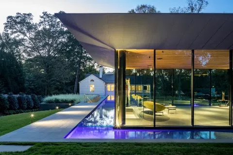 Modern glass house with illuminated pool and white traditional house in the background during dusk.