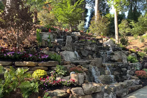 Natural stone waterfall surrounded by colorful flowers and greenery in a sunny garden setting.