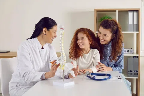 Pediatric doctor explaining spinal health to a smiling child and her mother in a clinic setting.