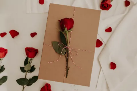 Brown gift box tied with red and white string and decorated with a single red rose on a white fabric background with rose petals.