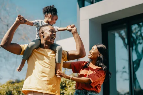 Happy family outdoors with father carrying daughter on shoulders and mother smiling nearby in sunny day.