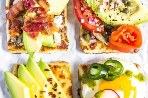 Four square toasts topped with avocado, bacon, tomato, egg, radish, jalapeno, and herbs on parchment paper.