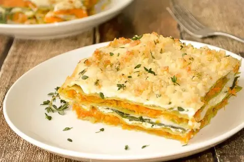 Slice of creamy vegetable lasagna topped with herbs and breadcrumbs on a white plate.
