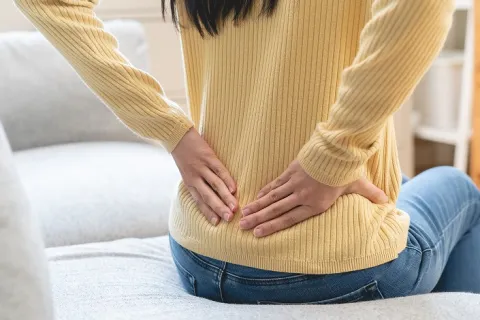 Person sitting on a couch holding their lower back, indicating back pain or discomfort indoors.