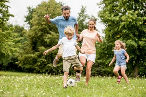 Family of four happily playing soccer together outdoors on a green field with trees in the background