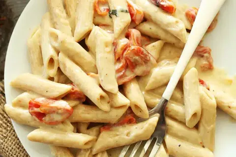Creamy penne pasta with cherry tomatoes on white plate with fork on wooden table.