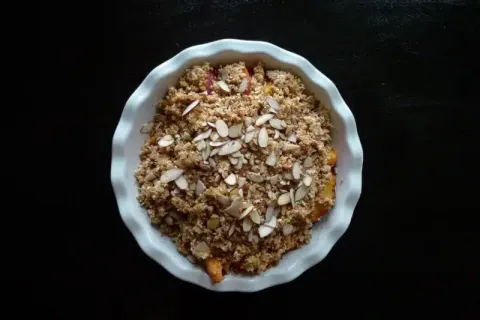 Top view of a fruit crumble dessert topped with sliced almonds in a white ceramic dish on black surface