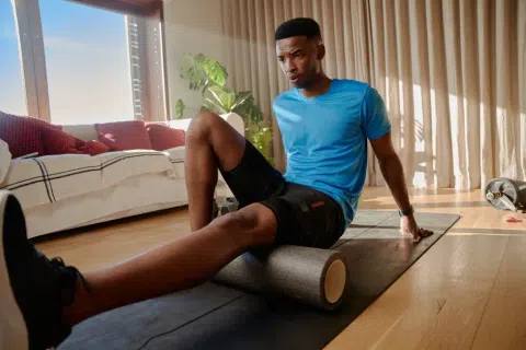 Man in blue shirt using foam roller on leg while exercising indoors on a yoga mat in bright living room