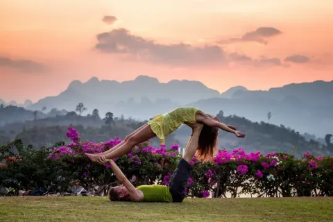 Couple practicing acro yoga outdoors at sunset with mountains and vibrant flowers in the background.