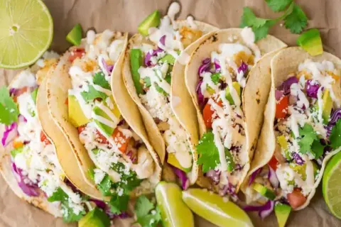 Fresh fish tacos with avocado, cabbage, cilantro, lime wedges, and creamy sauce on brown paper.
