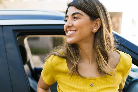 Smiling woman in yellow shirt leaning out of car window with urban background bright daylight.