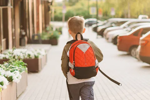 Young boy walking along city sidewalk with red backpack on sunny day near parked cars and planters.