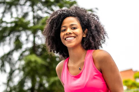 Smiling young woman in pink athletic wear outdoors with green trees and blurred background on a sunny day
