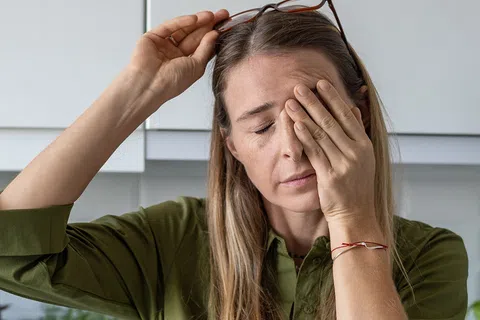 Woman with blonde hair in green shirt holding her forehead, appearing tired or stressed in a bright kitchen.