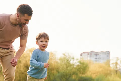 Father and young son running together in a sunny park with trees and distant buildings in the background