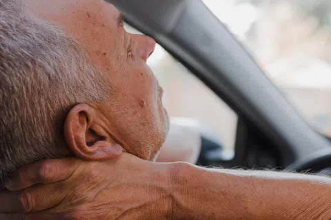 Senior man sitting in car holding neck with hand, showing discomfort or pain while driving.