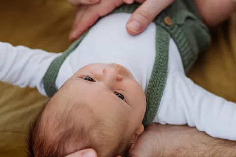 Baby lying on bed wearing white and green clothing, held gently by adults with blurred background.