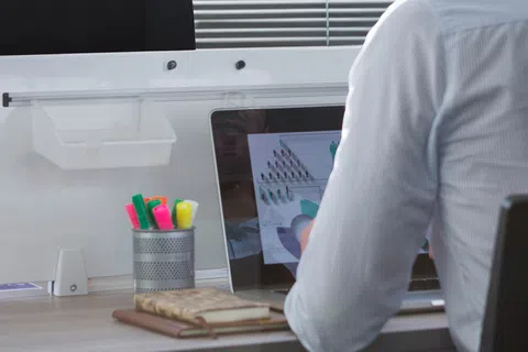 Person working on a laptop sitting in an ergonomic chair at a desk with office supplies, a notebook, and a modern lamp visible.