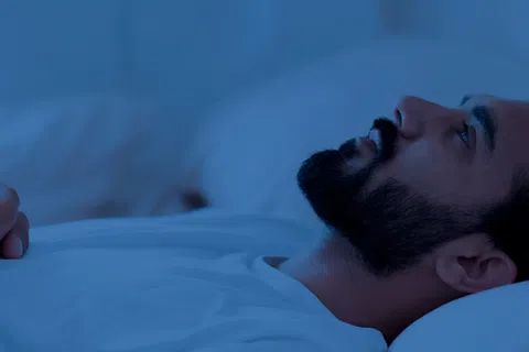 Man lying awake in bed with hands clasped, looking up, surrounded by a blue-toned dimly lit room.