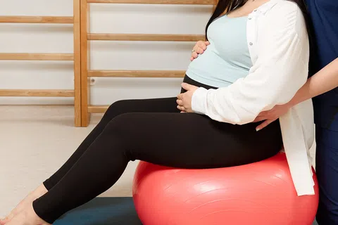 Pregnant woman sitting on exercise ball with therapist support in a physical therapy room.
