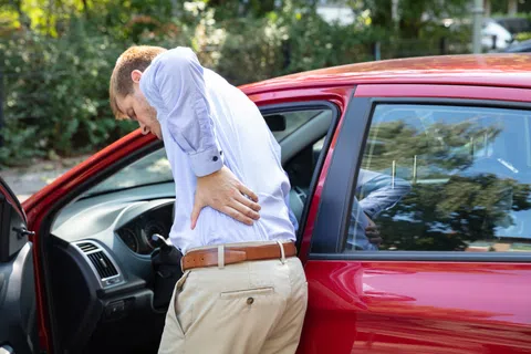 Man with back pain standing outside a red car with the driver door open in a sunny environment.