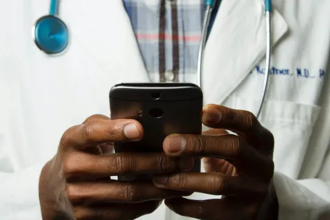 Close-up of a doctor wearing a white coat and stethoscope using a smartphone.