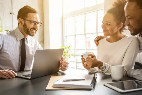 Happy couple consulting with a professional advisor at a sunny office, discussing documents and using a laptop.