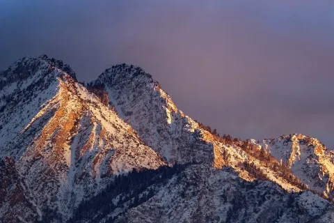 Snow-capped mountain peaks glowing with golden sunlight under a dramatic cloudy sky at sunset.