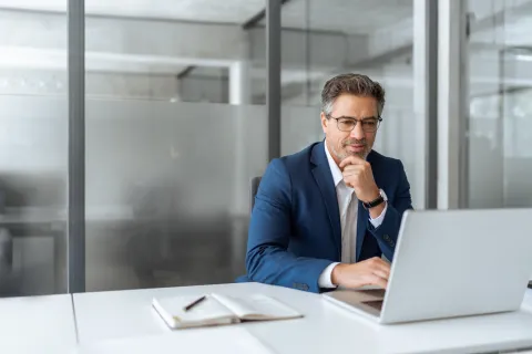 Businessman in blue suit working on laptop at desk in modern office with notebook and pen nearby.