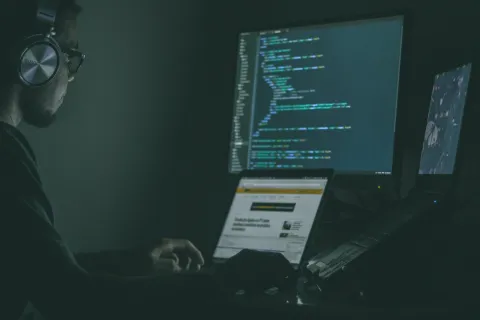 Person wearing headphones coding on multiple screens in a dimly lit room with programming code visible.