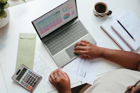 Person working on financial data using laptop, calculator, documents, and notebook on a white desk.