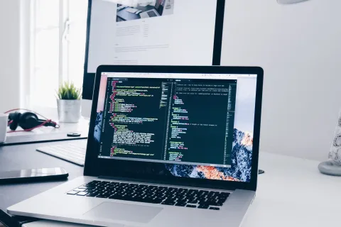 Laptop on desk displaying coding software in a modern bright office setup with headphones and coffee nearby
