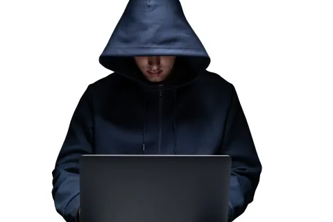 Person in dark hoodie using a laptop in low light with face partially hidden under hood.