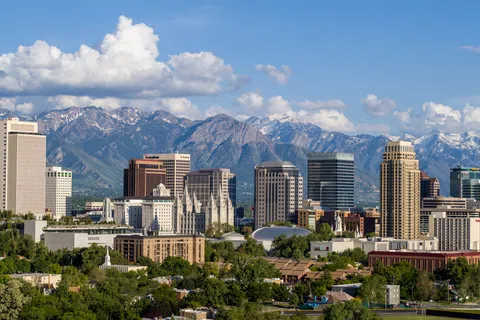 Salt Lake City skyline with modern buildings and snow-capped mountains under a blue sky with clouds.
