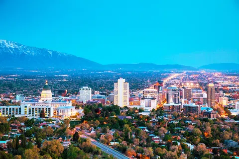Panoramic view of a city skyline at dusk with lit buildings, green trees, and snow-capped mountains in the background