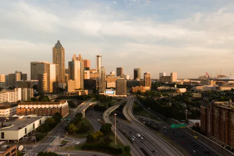 Aerial view of Atlanta skyline with highways, tall buildings, and a partly cloudy sky at sunset.