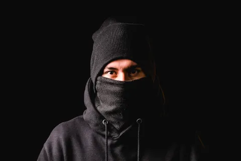 Man wearing black beanie and scarf covering his face against a dark background, showing intense eyes.
