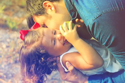 Father lovingly kisses smiling little daughter holding green apple outdoors in warm sunlight.