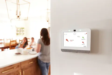 Smart home security system control panel mounted on a wall in a modern kitchen with family in background.