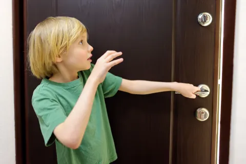 Young boy in green shirt reaching to open a dark wooden door with a silver handle and lock.