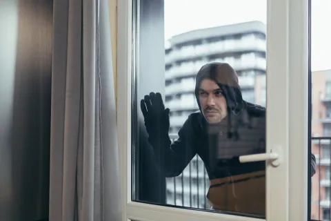 Man in black hoodie and gloves attempting to break into modern apartment through glass door at night.