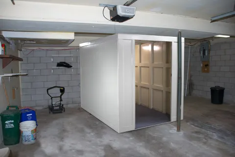 Basement room with concrete walls and floor containing a large white safe room with open door and various storage items