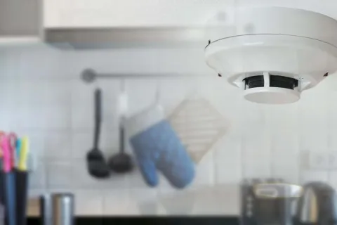 Close-up of a smoke detector installed on a kitchen ceiling with blurred utensils and oven mitts in the background