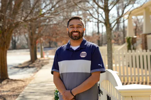 Smiling bearded man in blue-gray polo standing outdoors by a white picket fence on a sunny day.