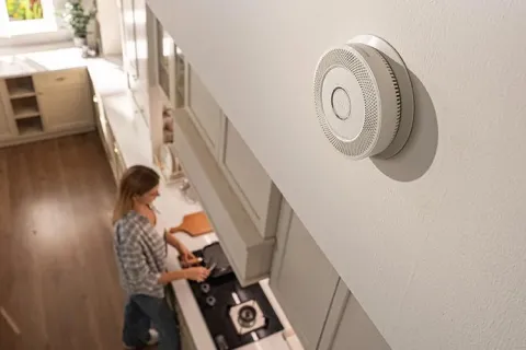 Modern smoke detector mounted on kitchen wall above a woman cooking in a light, spacious kitchen.