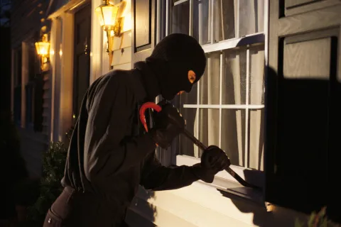 A masked burglar attempting to break into a house through a window at night.