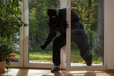 Burglar wearing black clothes and balaclava breaking into a house through a glass door with a crowbar.