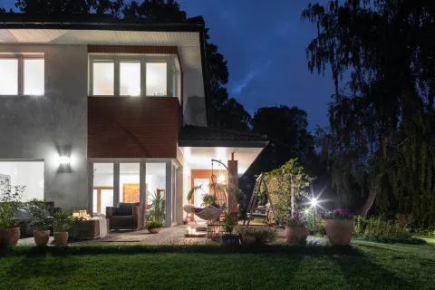 Modern two-story house with lit interior and outdoor patio featuring plants and cozy seating at night.